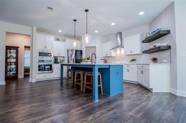 kitchen featuring hanging light fixtures, stainless steel appliances, white cabinetry, wall chimney exhaust hood, and an island with sink