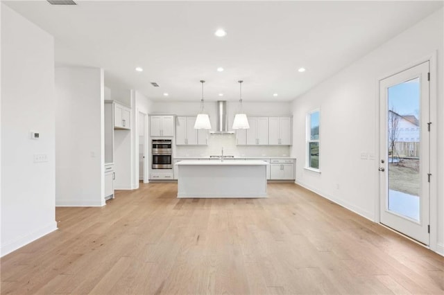 kitchen with tasteful backsplash, wall chimney range hood, light countertops, double oven, and light wood finished floors
