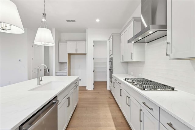 kitchen featuring light wood-style flooring, decorative light fixtures, wall chimney exhaust hood, stainless steel appliances, and a sink