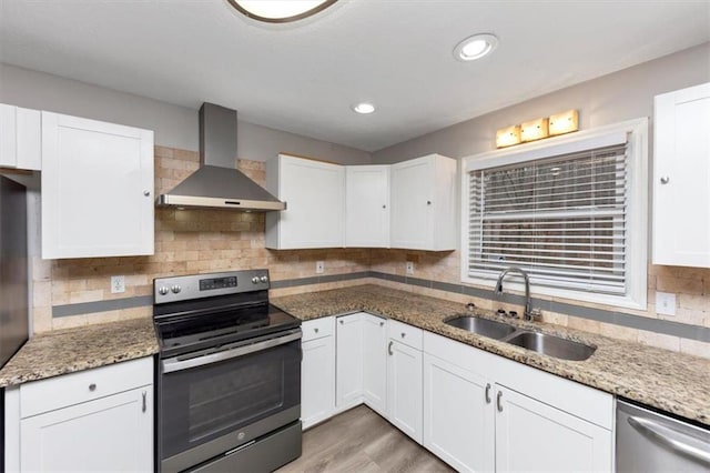 kitchen featuring wall chimney range hood, sink, stone counters, white cabinetry, and appliances with stainless steel finishes