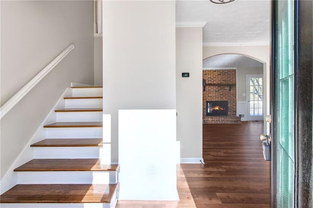 stairway featuring arched walkways, a fireplace, ornamental molding, wood finished floors, and baseboards