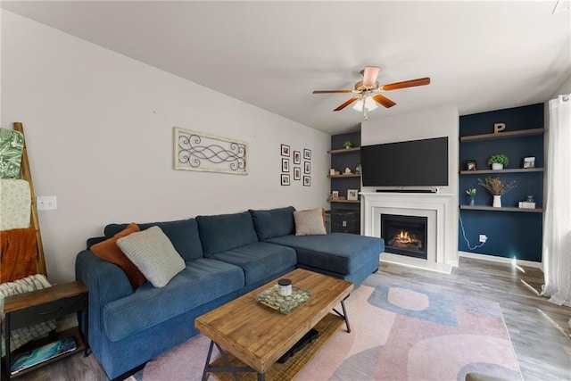 living room with wood finished floors, ceiling fan, a lit fireplace, and built in features