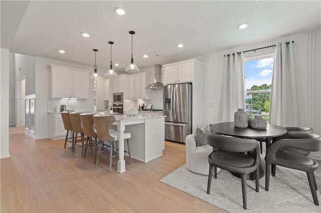 kitchen featuring white cabinets, an island with sink, hanging light fixtures, wall chimney range hood, and stainless steel appliances