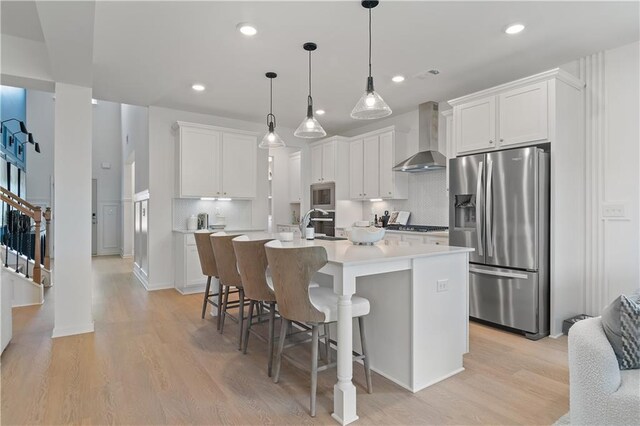 kitchen with an island with sink, appliances with stainless steel finishes, wall chimney exhaust hood, and white cabinetry