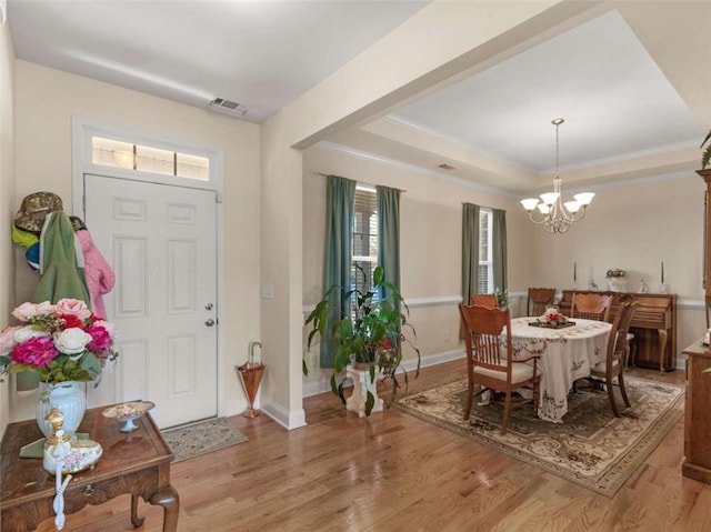 foyer entrance featuring a notable chandelier, a raised ceiling, visible vents, light wood-style floors, and ornamental molding