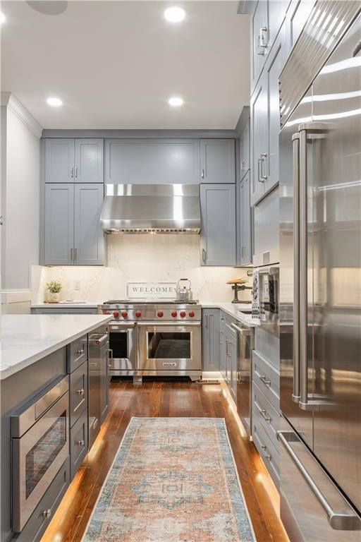 kitchen featuring tasteful backsplash, gray cabinets, built in appliances, dark wood-type flooring, and wall chimney exhaust hood