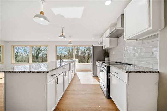 kitchen featuring light wood finished floors, tasteful backsplash, wall chimney exhaust hood, appliances with stainless steel finishes, and a kitchen island with sink