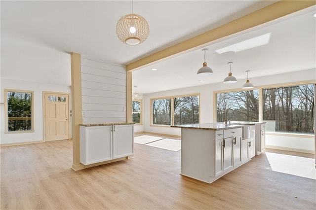 kitchen featuring white cabinets, light wood-style floors, an island with sink, decorative light fixtures, and stone counters