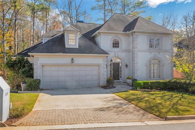 french country inspired facade with stucco siding, roof with shingles, a front yard, an attached garage, and concrete driveway