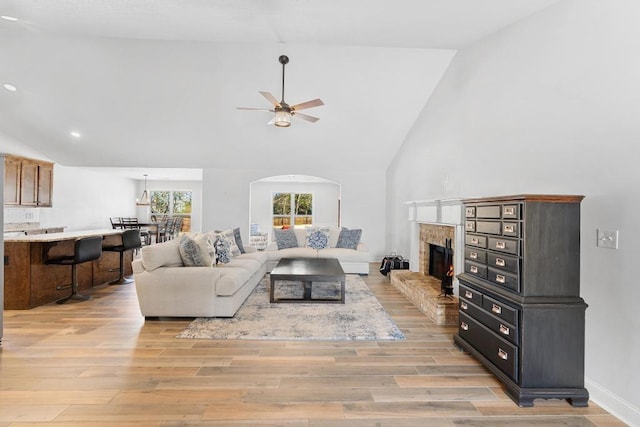 living area with light wood-type flooring, high vaulted ceiling, a tiled fireplace, arched walkways, and ceiling fan