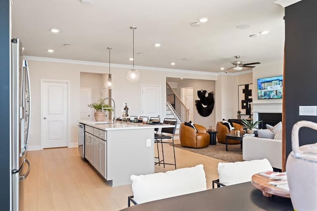 kitchen with an island with sink, ceiling fan, stainless steel appliances, decorative light fixtures, and light wood-type flooring