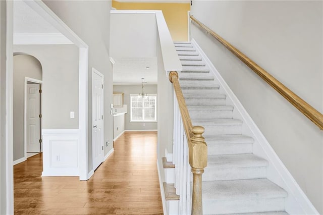 stairs with wood finished floors, an inviting chandelier, ornamental molding, wainscoting, and a decorative wall