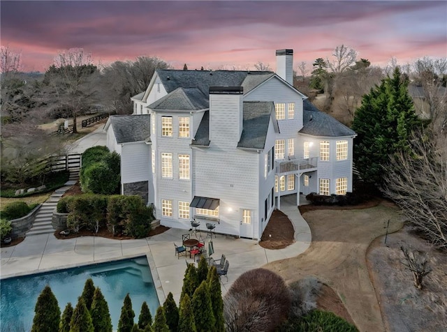 back of property at dusk featuring a chimney, fence, an outdoor pool, a balcony, and a patio area