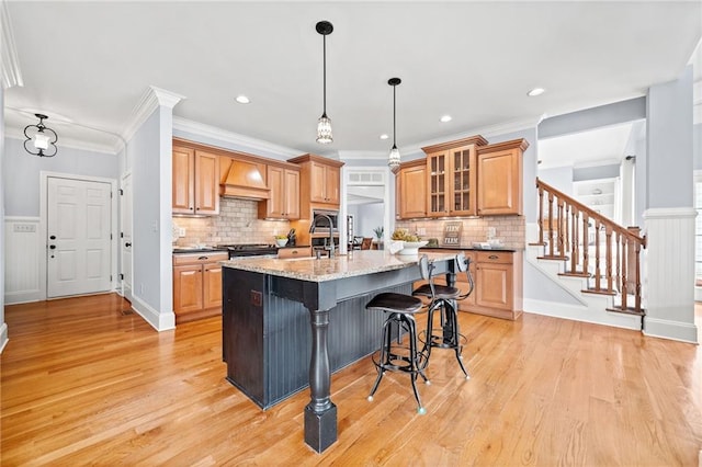 kitchen with light wood-style flooring, a center island with sink, backsplash, a kitchen bar, and custom range hood