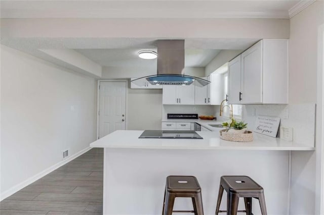 kitchen with kitchen peninsula, black electric stovetop, a kitchen breakfast bar, island range hood, and white cabinetry