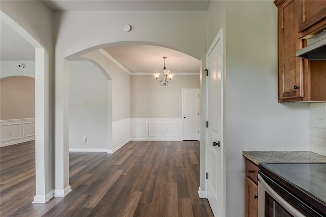 unfurnished dining area with dark wood-type flooring, a chandelier, crown molding, arched walkways, and a wainscoted wall