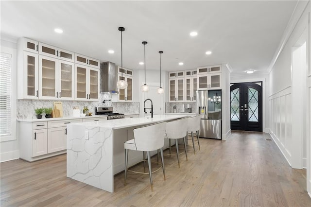 kitchen with a large island with sink, french doors, wall chimney range hood, white cabinetry, and stainless steel appliances