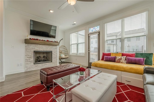 living area featuring wood finished floors, a ceiling fan, baseboards, ornamental molding, and a stone fireplace