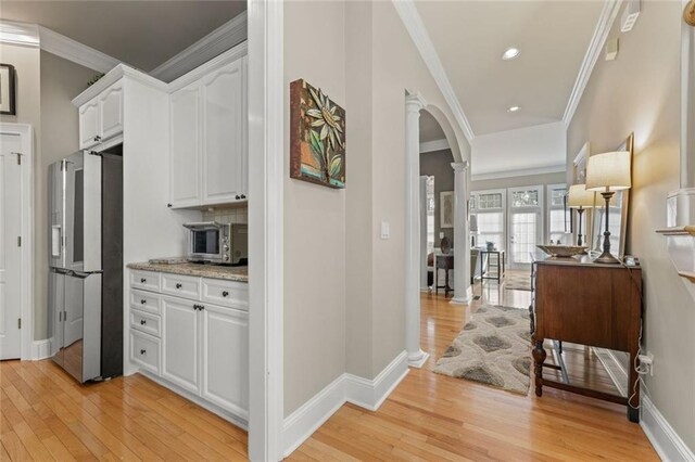 hallway featuring ornate columns, light wood-style flooring, crown molding, arched walkways, and baseboards