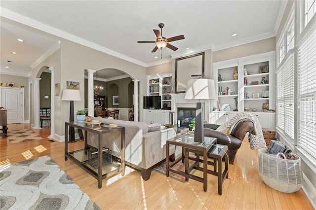 living room featuring arched walkways, decorative columns, ceiling fan, a glass covered fireplace, and light wood-style flooring