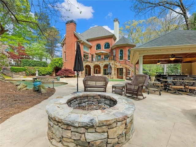 back of property with a balcony, a ceiling fan, an outdoor fire pit, a chimney, and a patio area