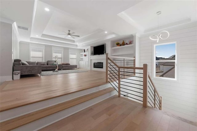 stairs featuring built in features, ceiling fan with notable chandelier, wood finished floors, a glass covered fireplace, and a raised ceiling