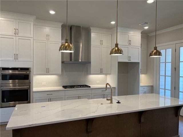 kitchen featuring hanging light fixtures, appliances with stainless steel finishes, a center island with sink, light stone countertops, and wall chimney range hood