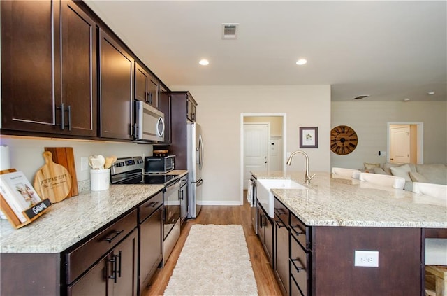 kitchen with dark brown cabinets, a center island with sink, light wood-type flooring, appliances with stainless steel finishes, and a sink