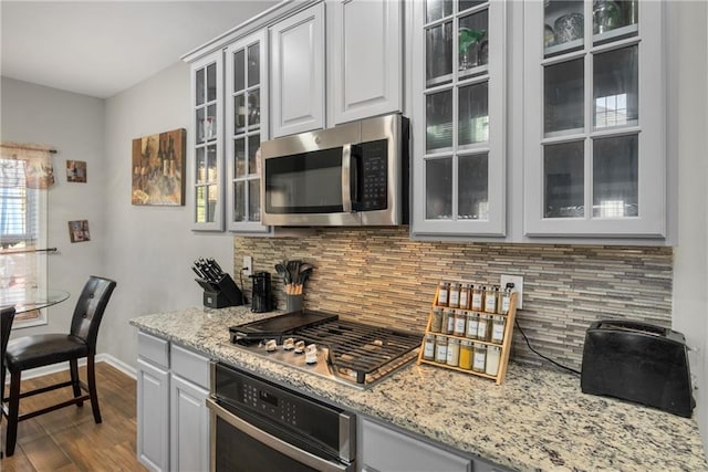 kitchen featuring light stone countertops, dark wood-style flooring, decorative backsplash, glass insert cabinets, and appliances with stainless steel finishes