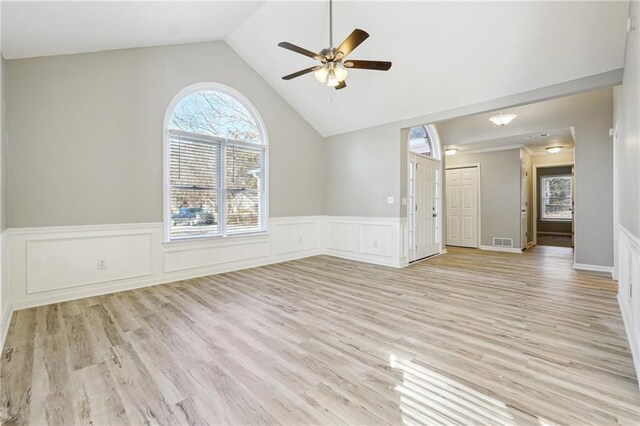 empty room with a wainscoted wall, lofted ceiling, visible vents, light wood-style flooring, and ceiling fan with notable chandelier