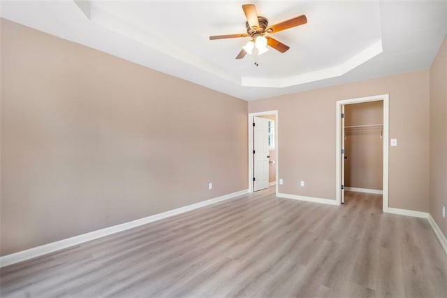 unfurnished bedroom featuring a closet, light wood finished floors, a tray ceiling, a walk in closet, and baseboards