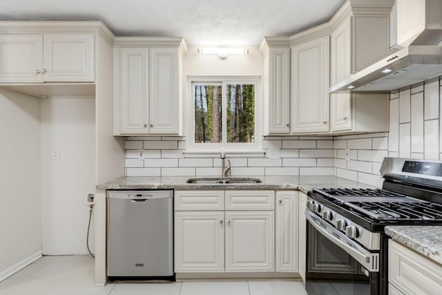 kitchen with wall chimney range hood, light stone counters, appliances with stainless steel finishes, and a sink