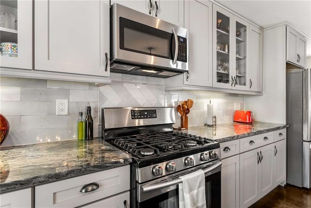 kitchen featuring stainless steel appliances, dark stone counters, glass insert cabinets, and backsplash