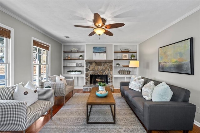 living room with ceiling fan, a stone fireplace, wood finished floors, visible vents, and crown molding