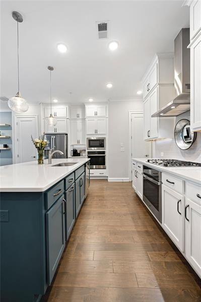 kitchen with a large island, hanging light fixtures, wall chimney range hood, white cabinets, and appliances with stainless steel finishes