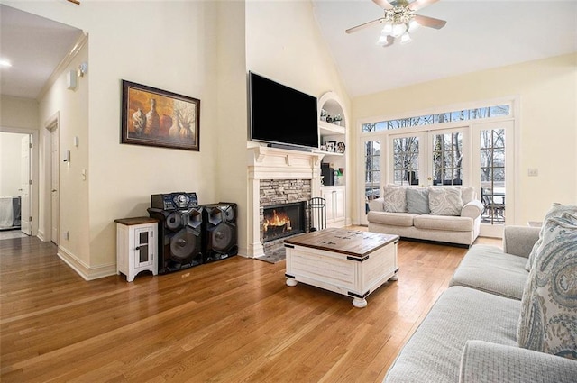 living area featuring built in features, a ceiling fan, light wood finished floors, high vaulted ceiling, and a stone fireplace