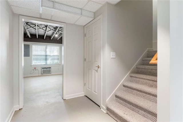 stairway featuring concrete flooring, baseboards, a drop ceiling, a wall mounted AC, and electric panel