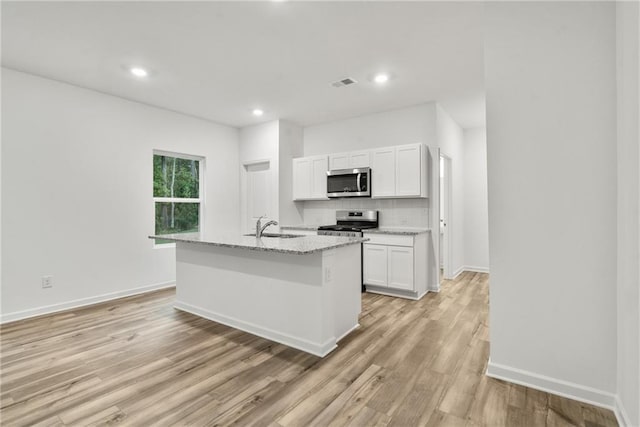 kitchen with stainless steel appliances, white cabinetry, light stone countertops, and a kitchen island with sink