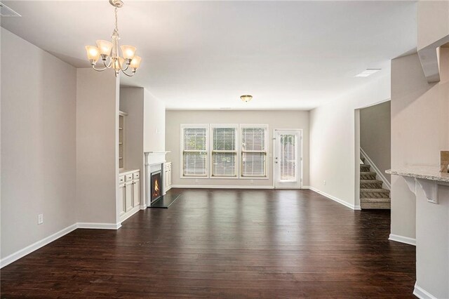 unfurnished living room featuring a warm lit fireplace, baseboards, dark wood-style floors, an inviting chandelier, and stairs