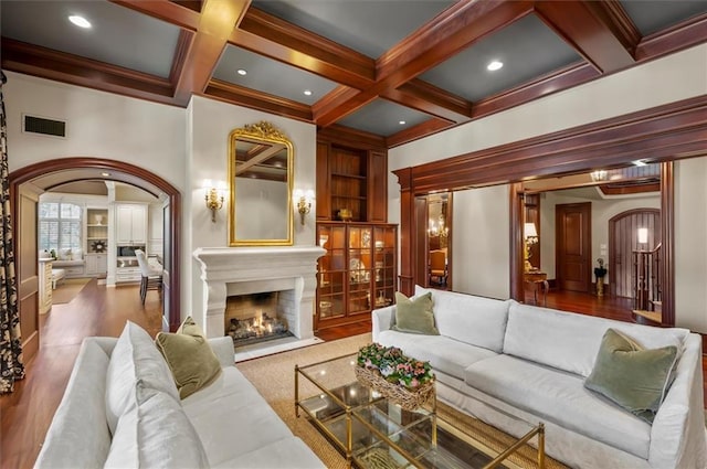 living area featuring arched walkways, beamed ceiling, a warm lit fireplace, coffered ceiling, and built in features