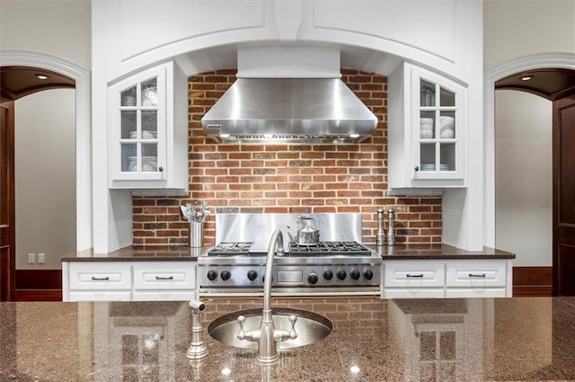 kitchen with wall chimney exhaust hood, glass insert cabinets, dark stone counters, and tasteful backsplash