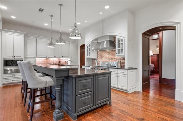 kitchen featuring arched walkways, range, wall chimney exhaust hood, a warming drawer, and white cabinets