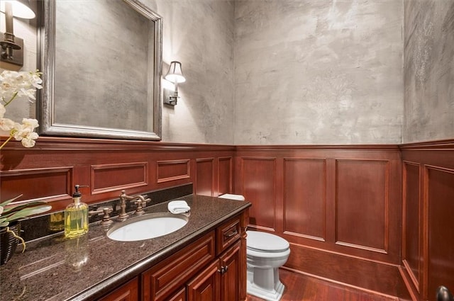 bathroom with toilet, wood finished floors, vanity, a decorative wall, and wainscoting