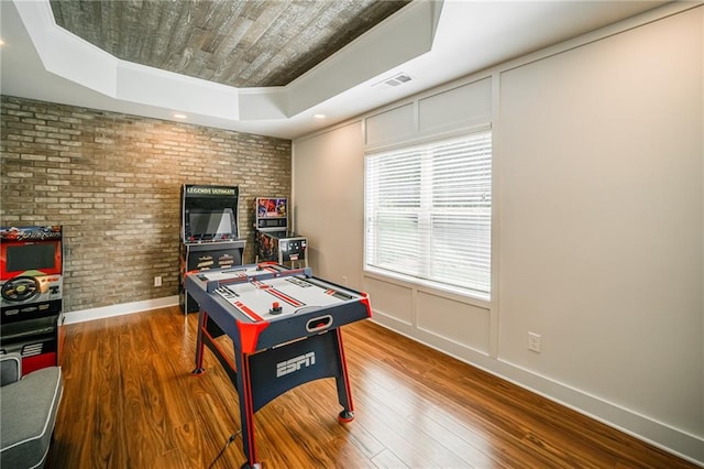 recreation room with brick wall, hardwood / wood-style flooring, crown molding, and a raised ceiling