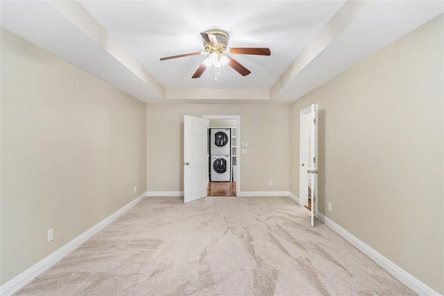 empty room with ceiling fan, light carpet, stacked washer / dryer, baseboards, and a tray ceiling