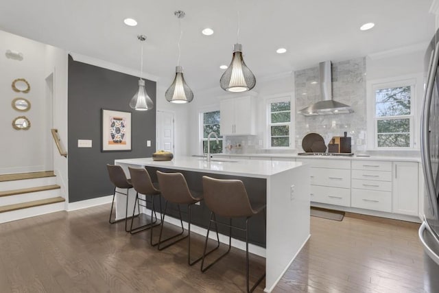 kitchen featuring wall chimney range hood, sink, white cabinetry, backsplash, and a center island with sink