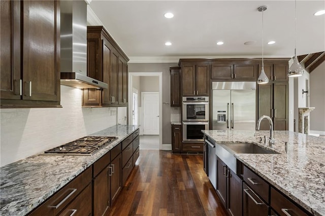 kitchen with appliances with stainless steel finishes, wall chimney range hood, a sink, crown molding, and dark wood finished floors
