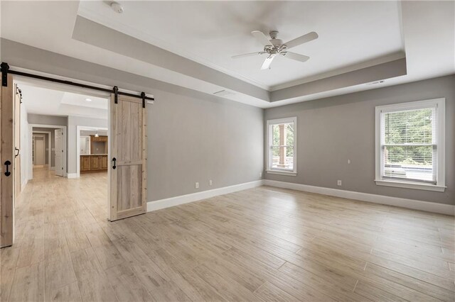 empty room with a raised ceiling, a barn door, crown molding, a ceiling fan, and baseboards