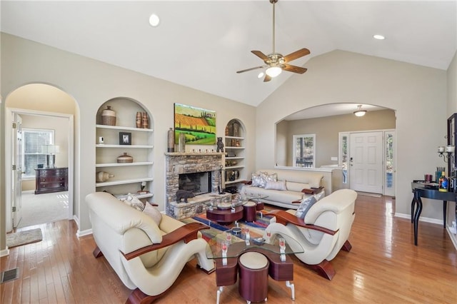 living room featuring built in shelves, a stone fireplace, vaulted ceiling, ceiling fan, and light hardwood / wood-style flooring