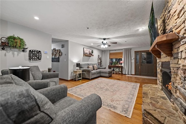 living room featuring ceiling fan, a textured ceiling, a stone fireplace, light wood-style floors, and recessed lighting